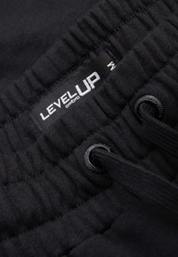 Gros plan sur une ceinture élastique noire avec une étiquette « LEVEL UP ombre » et des œillets noirs pour cordon sur un tissu texturé.
