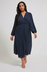 Navy blue pleated dress with a wrap V-neck, long sleeves, and dotted texture. Sheer side panels and a flowing silhouette. Strappy silver heels.