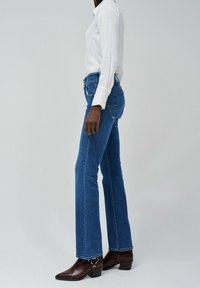 Niebieskie dżinsy w kształcie dzwonów z średnim stanem, zestawione z białą bluzką z długim rękawem i brązowymi botkami za kostkę z metalicznym akcentem.