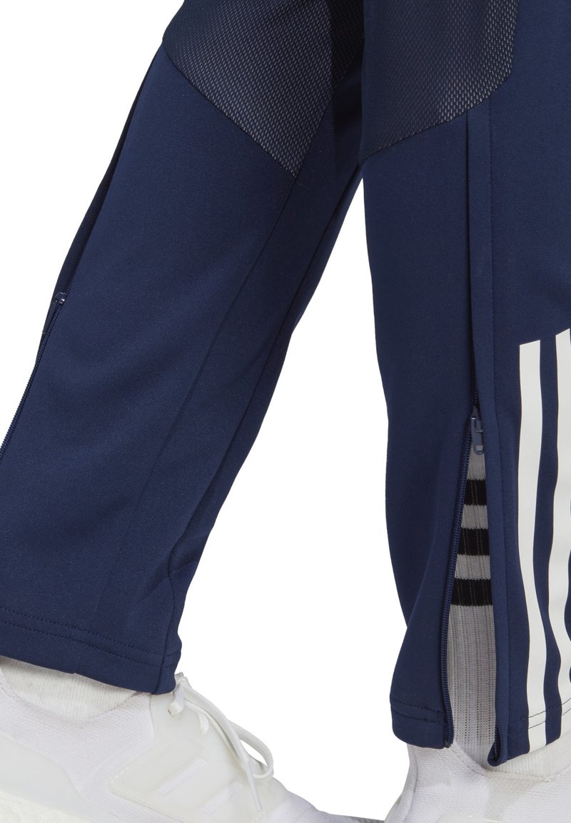 Pantalons de sport navy avec des détails en mesh, dotés d'une fermeture éclair sur le bas de la jambe et de rayures verticales blanches. Tissu texturé et finition lisse.
