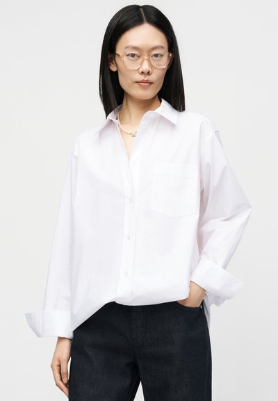 Femme aux cheveux noirs et raides portant des lunettes à monture transparente, une chemise blanche oversize boutonnée, un pantalon bleu marine et un collier de perles.