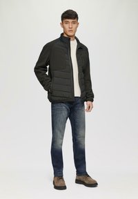 Schwarze Pufferjacke mit hohem Kragen, strukturierten Schulterpatches und einem Reißverschluss vorne. Kombiniert mit blauen Jeans und braunen Schuhen, stehendem Modell.