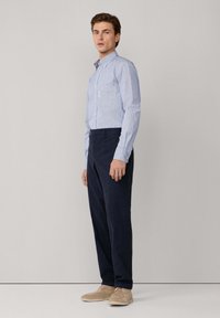 Uomo che indossa una camicia a righe blu chiaro con bottoni, pantaloni blu navy scuri e scarpe in suede beige, in piedi contro uno sfondo grigio chiaro semplice.