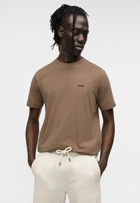 T-shirt en coton marron avec un col rond et des manches courtes, arborant un petit logo noir "BOSS" sur la poitrine. Associé à un pantalon couleur crème.