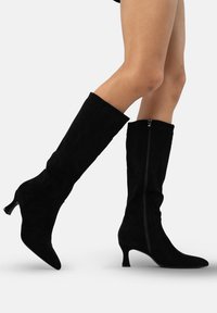Botas altas de ante negro hasta la rodilla con punta afilada, un tacón de kitten delgado y un detalle de cremallera lateral para facilitar el uso. Textura suave en toda la bota.