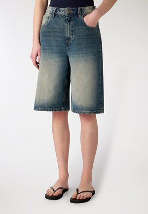 Donna che indossa pantaloncini Bermuda in denim blu sbiadito, canottiera nera e infradito nere, in piedi con una mano in tasca.