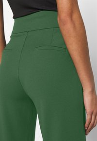 High-waisted green trousers with a smooth, textured fabric. Features two back pockets and a sleek, form-fitting silhouette.