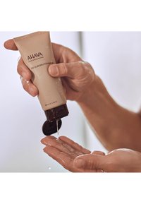 Gel esfoliante per la pulizia AHAVA in un tubo beige con un cappuccio nero, versa un gel trasparente su una mano bagnata, mostrando una texture liscia e lucente.