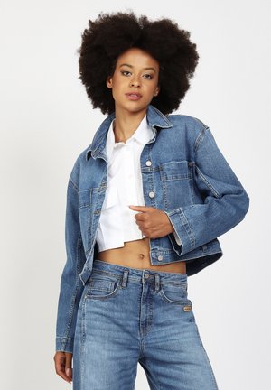 Junge Frau mit Afro-Haar, die ein bauchfreies weißes Hemd, eine blaue Jeansjacke und hoch taillierte blaue Jeans vor einem schlichten Hintergrund trägt.
