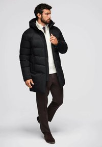 Giacca a piumino nera con cappuccio, dotata di chiusura a zip, design trapuntato e due tasche frontali, indossata sopra un maglione bianco e pantaloni marroni.