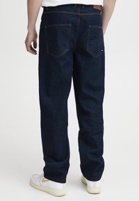 Vaqueros de mezclilla azul oscuro con un corte relajado, que cuentan con doble bolsillo trasero y costuras contrastantes visibles. Se llevan con zapatillas blancas.
