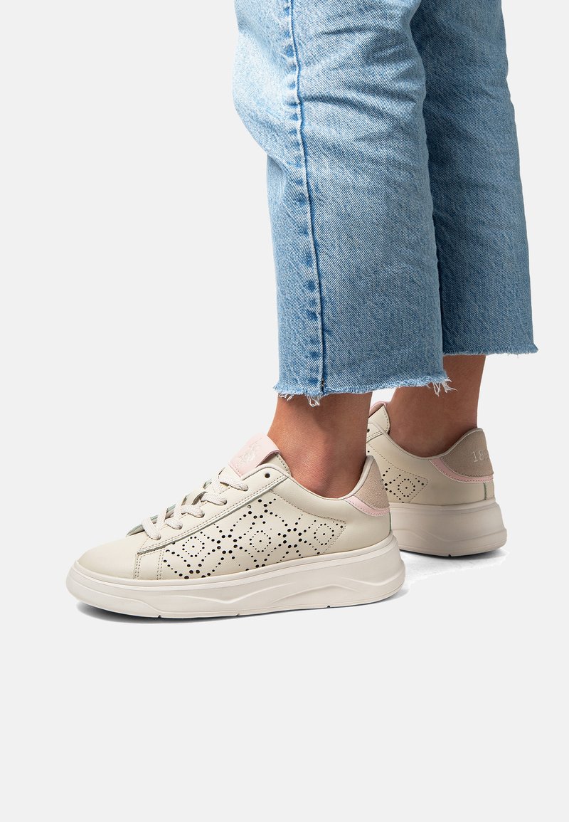 Beiga perforerade sneakers med en slät finish, utrustade med en ljusrosa bakaccent och vit plattformssula. Avsmalnande jeans med uppvikta ben synliga.