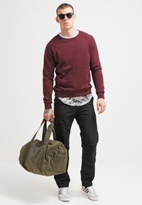 Sweatshirt bordeaux, pantalon cargo noir, baskets grises et lunettes de soleil sombres. Portant un grand sac de sport vert olive avec un imprimé graphique.