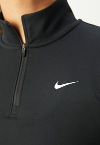 Musta urheilullinen vetoketjullinen paita, jossa on korkea kaulus, teksturoitu kangas ja suuri valkoinen Nike-swoosh-logo rinnassa.