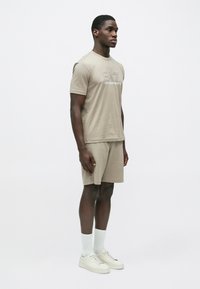 Άνδρας μοντέλο όρθιος, φοράει μπεζ t-shirt EA7 Emporio Armani και ταιριαστά σορτς με λευκές κάλτσες και λευκά αθλητικά παπούτσια, μπροστά σε απλό φόντο.