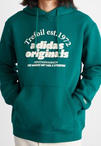 Grön hoodie med dragsko huva, stor vit textdesign och framficka. Har Adidas-logotyp på ärmsluten och ribbade fållar.
