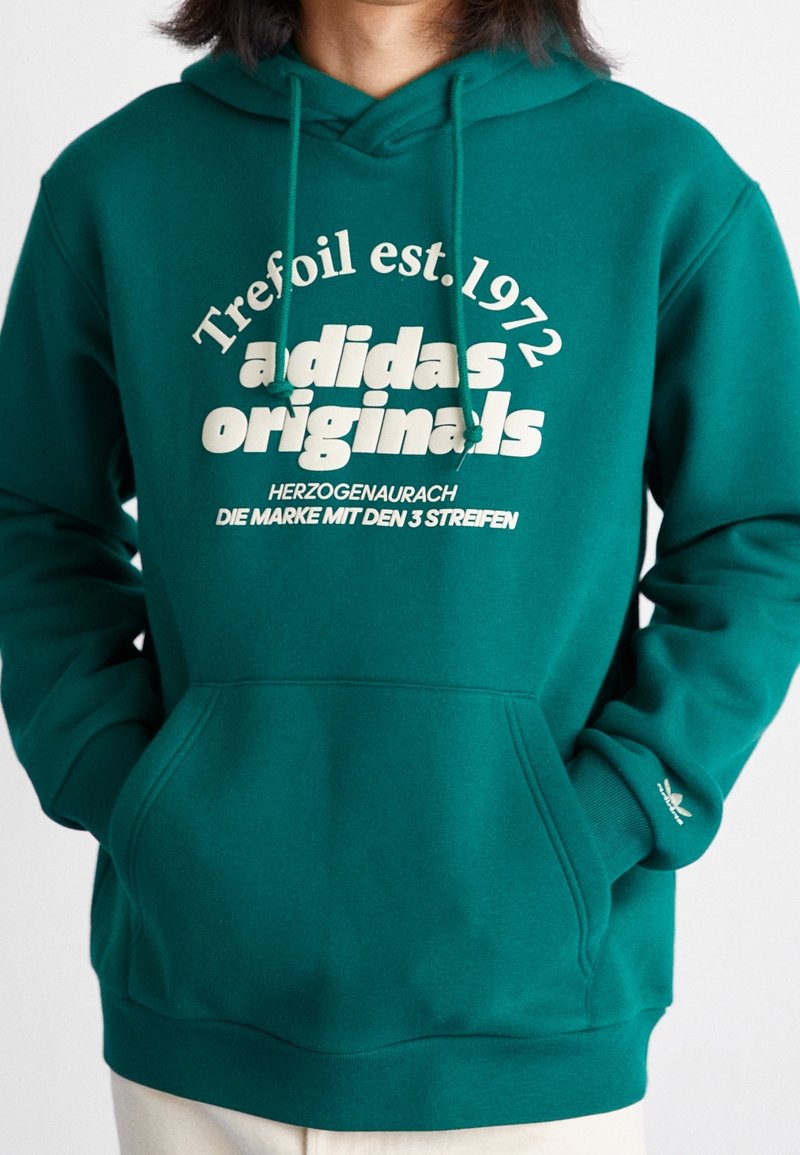 Grön hoodie med dragsko huva, stor vit textdesign och framficka. Har Adidas-logotyp på ärmsluten och ribbade fållar.