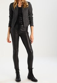 Femme portant un pantalon en cuir noir à lacets, des bottes noires à lacets, un haut noir avec détail corset, et une veste grise texturée sur fond blanc.
