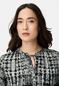 Jeune femme aux cheveux ondulés foncés portant un chemisier à motifs noir et blanc avec un col noué à l'avant, face à l'avant avec une expression neutre.