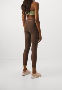 Leggings atletiche marroni con vita alta e pannelli testurizzati, abbinate a scarpe sportive rosa e una maglietta verde chiaro.