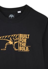 T-shirt en coton noir avec un graphisme jaune représentant un dinosaure et le texte "CONÇU POUR LES AUDACIEUX" avec le logo Timberland en dessous.
