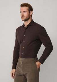Hombre con barba, vistiendo una camisa de botones de manga larga marrón oscuro y pantalones marrones, de pie con una mano en el bolsillo frente a un fondo simple.