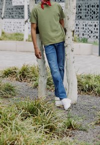 Camiseta de manga corta verde oliva, vaqueros azules, zapatillas blancas y un pañuelo rojo. Persona apoyada contra un árbol pálido en un área de hierba.