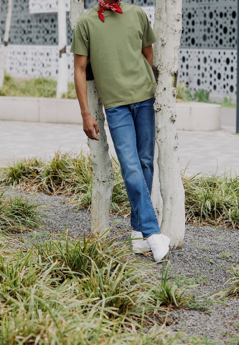 Camiseta de manga corta verde oliva, vaqueros azules, zapatillas blancas y un pañuelo rojo. Persona apoyada contra un árbol pálido en un área de hierba.