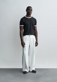 Mann steht vor einer schlichten Wand und trägt ein schwarzes T-Shirt mit weißer Einfassung, weiße, weit geschnittene Hosen, einen schwarzen Gürtel und schwarze Schuhe.