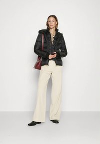 Svart vadderad jacka med dragkedja framtill och huva, med en texturerad design. Modellen matchade den med beige byxor med vida ben och en vinröd handväska.