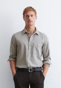 Helles graues Hemd mit Knopfleiste und Tasche, langärmlig mit hochgerollten Ärmeln, aus strukturiertem Stoff. Getragen mit schwarzen Hosen und einem Gürtel.