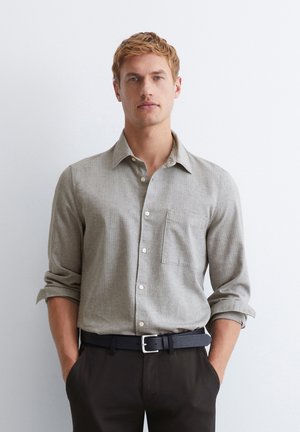 Helles graues Hemd mit Knopfleiste und Tasche, langärmlig mit hochgerollten Ärmeln, aus strukturiertem Stoff. Getragen mit schwarzen Hosen und einem Gürtel.