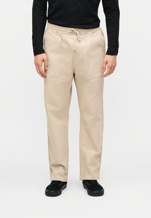 Pantalones beige con cordón y grandes bolsillos delanteros, combinados con una camiseta negra de manga larga y zapatillas negras, mostrados desde la cintura hasta los pies.