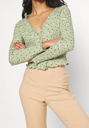 Kobieta ze skrzyżowanymi ramionami, ubrana w zielony, kwiecisty sweter na guziki oraz beżowe spodnie z wysokim stanem na jednolitym tle.