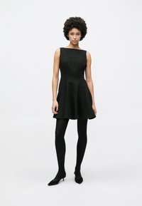 Μαύρο αμάνικο φόρεμα με εφαρμοστή κορσέ και φαρδύ φούστα, με λεία υφή. Φορεμένο με μαύρες κάλτσες και παπούτσια με μυτερή μύτη.