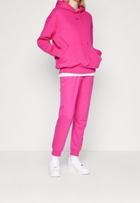 Pink hoodie with a front pocket and adjustable drawstring, paired with matching pink joggers. White sneakers complete the outfit.