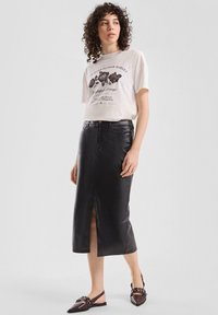 Schwarzer Ledermidirock mit einem vorderen Schlitz, kombiniert mit einem weißen T-Shirt mit Grafik. Auffällige Details sind ein Knopfverschluss und Gürtelschlaufen.