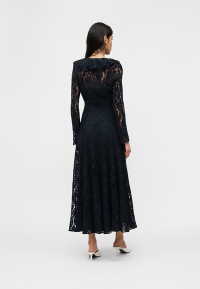 Sieviete, tērpusies garā, caurspīdīgā melnās mežģīņu kleitā ar garām piedurknēm un volānu apkakli, stāvot ar muguru, valkājot baltas papēžus ar atvērtu papēdi.