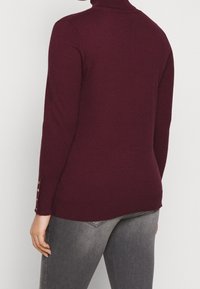 Pull col roulé bordeaux à manches longues, avec une coupe ajustée et trois boutons décoratifs aux poignets. Texture tricotée douce.