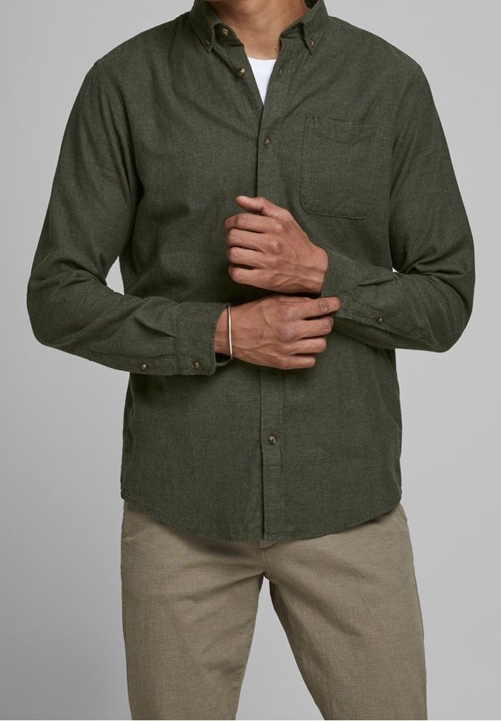 Man adjusting cuff of dark green button-up shirt, wearing beige pants and a silver bracelet on left wrist, against plain gray background.