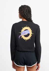 Maglietta cropped nera a maniche lunghe con una grafica di un serpente colorato circondata da un design di sole dorato sul retro. Tessuto dalla texture liscia.