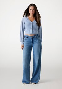 Svetlo modri pleteni kardigan z V-izrezom in gumbi, v kombinaciji s širokimi modrimi jeans hlačami. Obleka je izdelana iz teksturiranega materiala in ima krajšan dizajn.