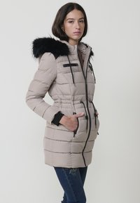 Chaqueta acolchada beige con capucha de pelo sintético negro, bolsillos con cremallera, cintura elástica y detalles de costura horizontal. Tejido cálido y ligero.