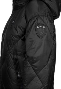 Giacca a vento nera con finitura testurizzata, dotata di cappuccio, tasche con zip e una toppa rotonda nera con il logo "Blauer" sulla spalla.