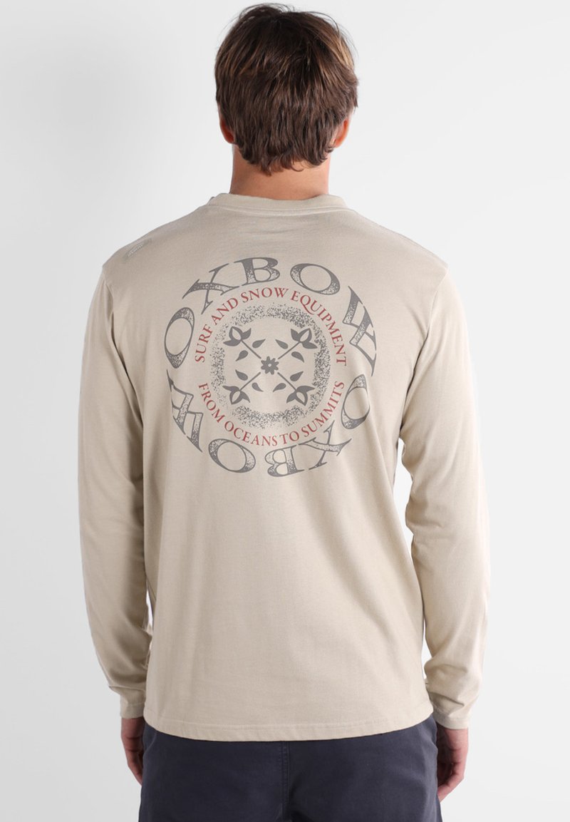T-shirt à manches longues beige clair avec un design graphique circulaire au dos, comportant du texte en gris et rouge sur un tissu en coton.