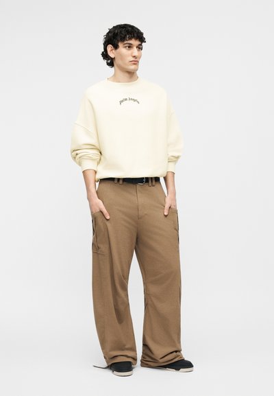 Νεαρός με σγουρά μαλλιά φορεί εκρού φούτερ με το κείμενο «palm angels», χαλαρό καφέ παντελόνι cargo, μαύρη ζώνη και μαύρα αθλητικά παπούτσια.