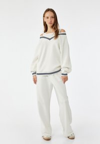 Biała bluza off-shoulder z granatowymi paskami, zestawiona z dopasowanymi białymi luźnymi spodniami. Miękki materiał i swobodny krój.