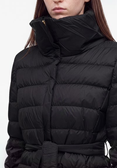 Femme portant une doudoune noire matelassée avec un col haut et une ceinture, montrée des épaules aux lèvres sur fond blanc.