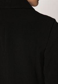 Černý kabát s hladkou texturou a strukturovaným designem, který má upravený límec a čisté linie podél zad.