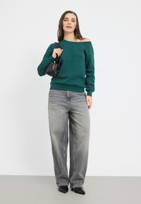 Sudadera verde sin hombros, vaqueros gris de pierna ancha, bolso negro y tacones negros de punta. Textura suave, diseño informal, corte moderno.